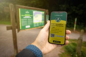 Person holding their mobile phone outwards showing the HiiKER App as they stand in front of a forest park sign